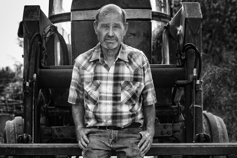 Older man in plaid shirt sitting on the back of a truck, black and white portrait.