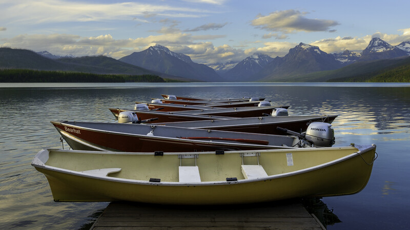 Yellow rowboat in foreground with brown wooden boats lined up behind it on calm water, snow-capped mountains in distance under partly cloudy sky.