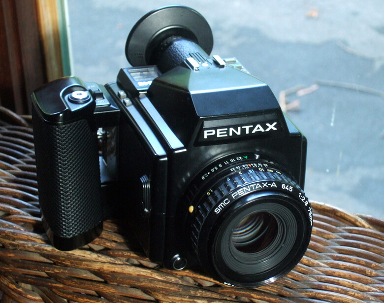 Pentax 67 medium format camera with lens mounted on wooden surface near water.