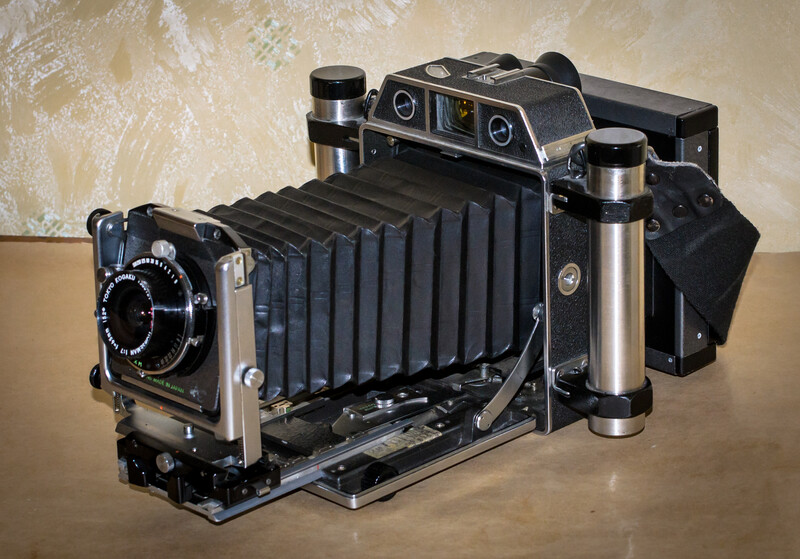 A vintage large format film camera with accordion-style bellows and dual lens assemblies mounted on a textured beige background.