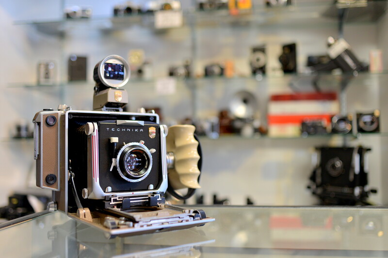 Vintage large-format camera displayed on a table with blurred collection of cameras and photography equipment in the background.