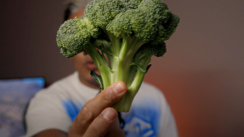 Child's hand holding a fresh broccoli floret with visible stems against a blurred indoor background.
