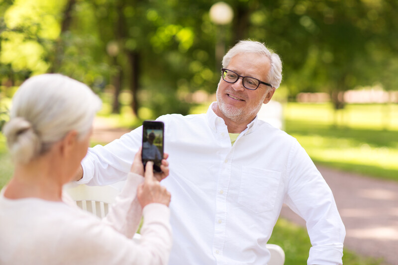 Older woman photographing a smiling man in glasses outdoors using a smartphone.