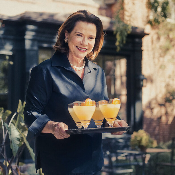 Woman in black jacket holding a tray of three yellow cocktails in an outdoor courtyard setting.