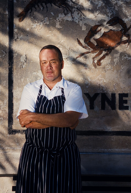 Chef in dark pinstriped apron and white shirt standing against weathered wall with shadow overlay.