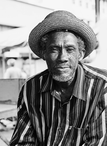 Black and white portrait of an older man wearing a striped shirt and straw hat, looking directly at camera.