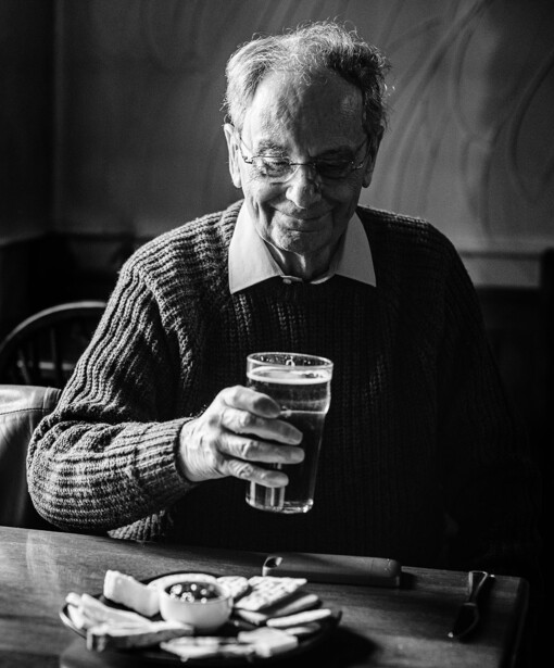 Older man in striped cardigan holding a glass of beer at a table with food and cigarettes.