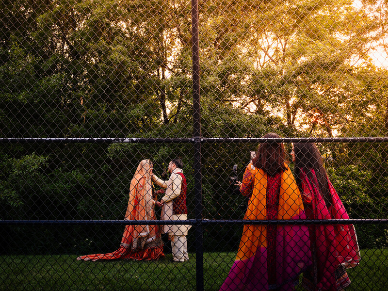Women in vibrant traditional clothing gathered behind a chain-link fence at golden hour.