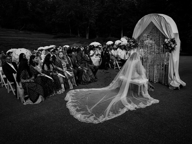 Bride in flowing veil walking down outdoor wedding aisle with guests seated on both sides.