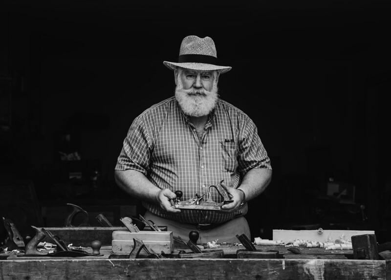 Bearded man in checkered shirt and fedora operating a large format camera or view camera at a workstation.