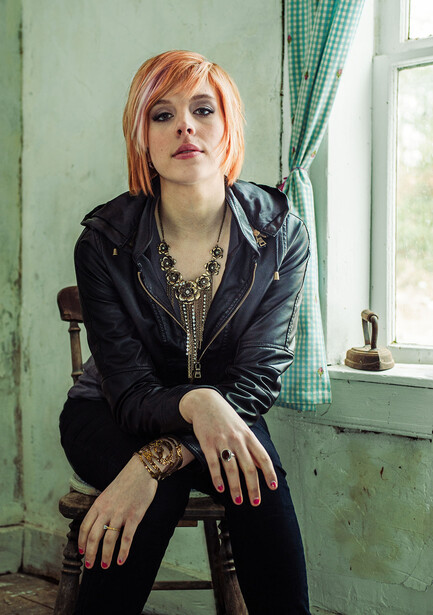Person with reddish hair sitting on a chair against a weathered turquoise wall with a window.