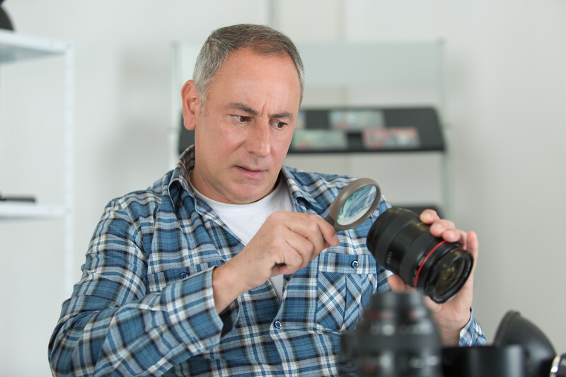 Photographer in blue plaid shirt examining a camera lens in a bright studio setting.