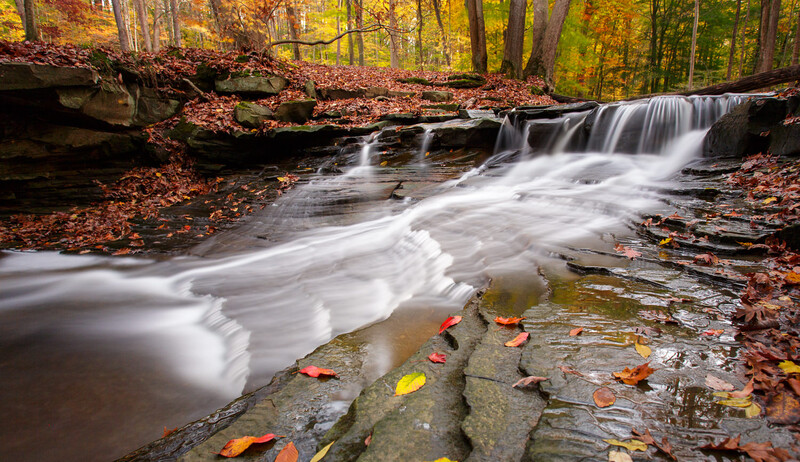 Long-exposure waterfall cascading through autumn forest with fallen leaves on rocks in foreground.