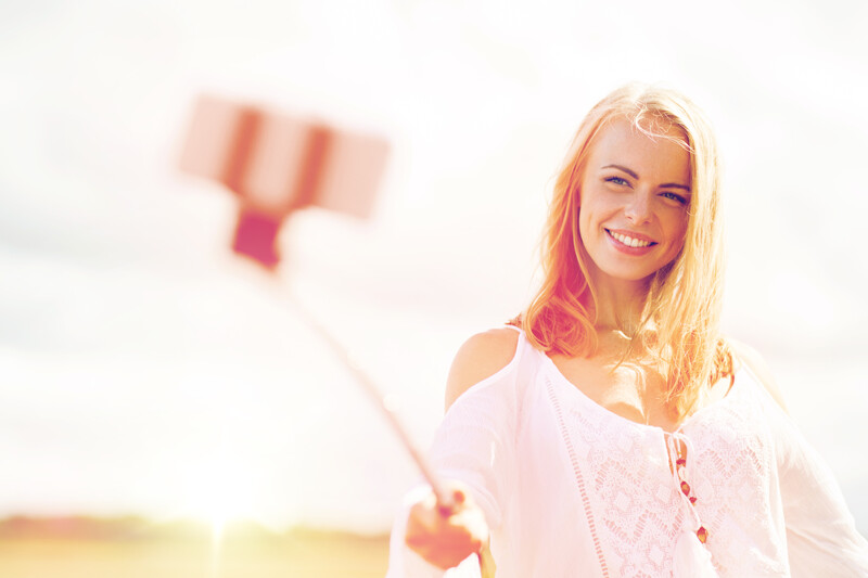 Smiling woman with blonde hair holding a selfie stick with a blurred smartphone attachment.