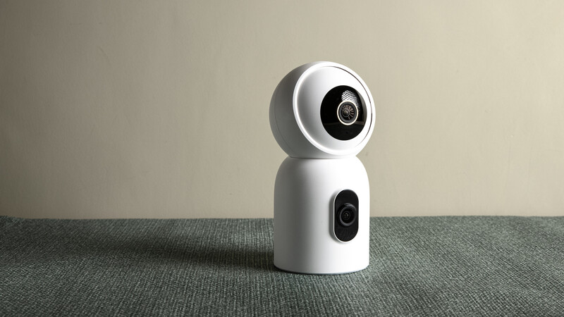 White spherical security camera with dual lens modules mounted on gray fabric surface against neutral background.