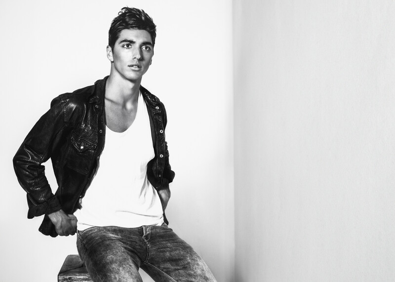 Man in black leather jacket and white shirt seated against white background.