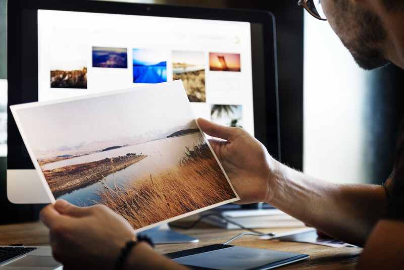 Photographer reviewing landscape photograph prints while viewing photo gallery on desktop monitor.