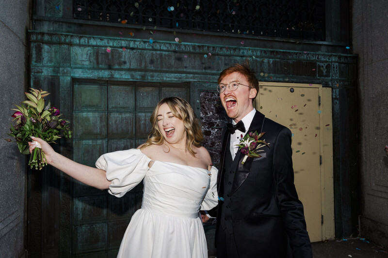 Bride and groom celebrating joyfully in front of a weathered teal door, bride holding bouquet aloft.