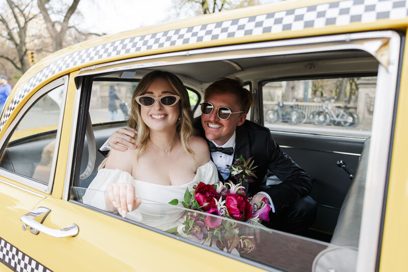 Bride and groom smiling through the window of a yellow vintage taxi decorated with a checkered pattern.