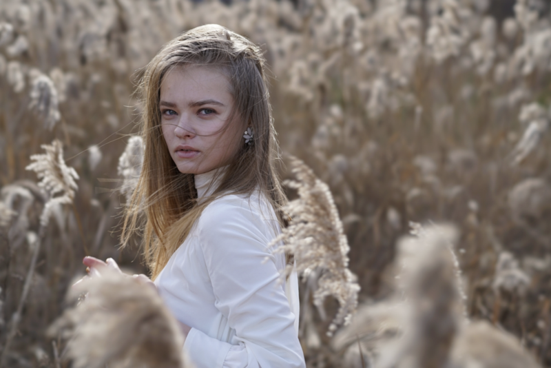 Young girl with long blonde hair in white shirt, standing in dried field looking at camera.