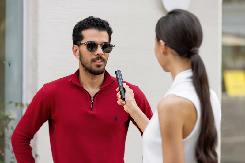 Man in red pullover wearing sunglasses being interviewed outdoors by woman holding microphone.