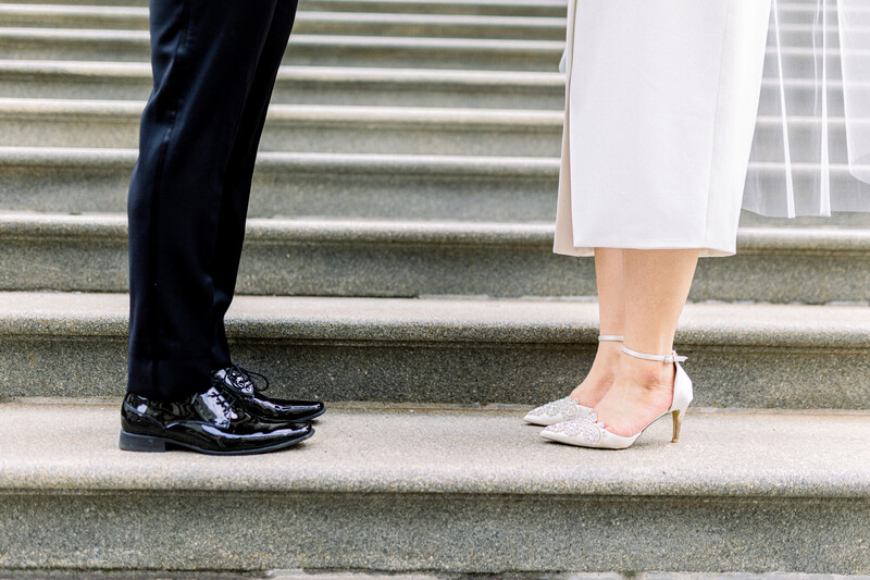 Close-up of two people's feet on stone steps, one wearing black dress shoes and dark trousers, the other in white heeled sandals and a light skirt.