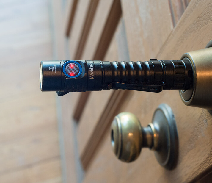 Close-up of a tactical flashlight with ribbed grip and red lens filter mounted on a rifle, with blurred wooden furniture background.