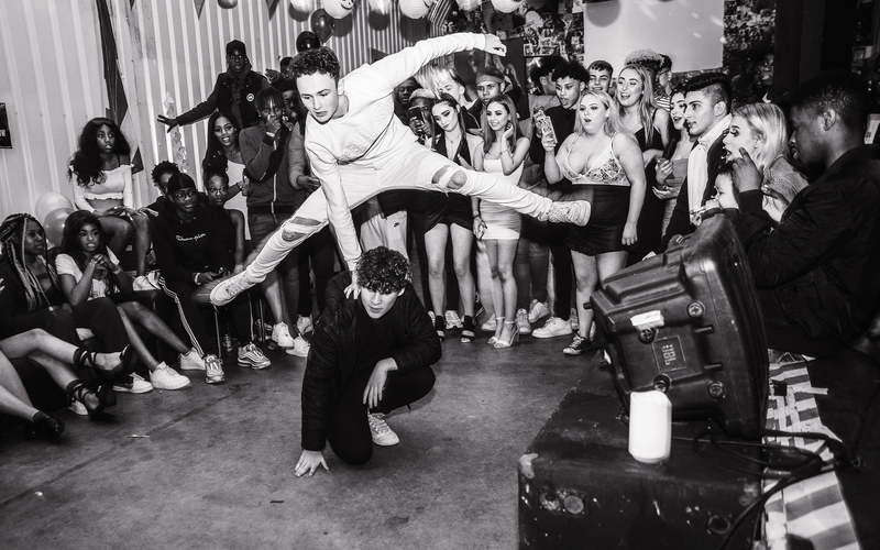 Breakdancer mid-flip in a packed indoor cypher with spectators circling the performance space.