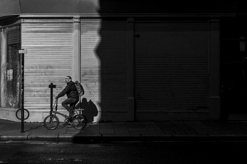 Person sitting on a small bike against a corrugated metal wall, lit by dramatic diagonal shadows from a window frame.