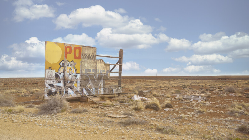 Abandoned roadside sign structure in desert landscape under blue sky with clouds.