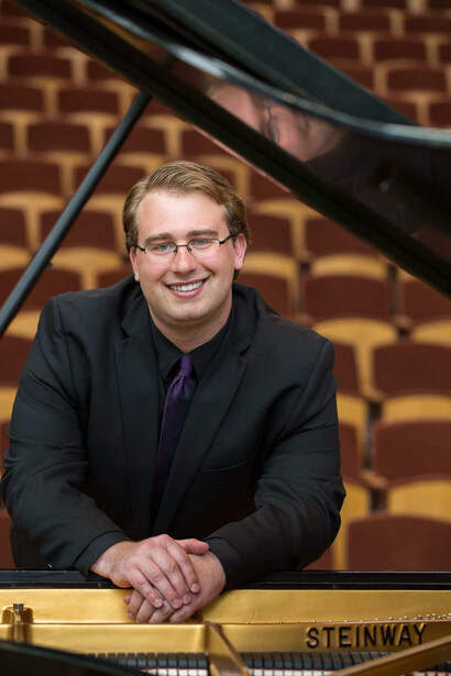 Man in dark blazer and glasses smiling at camera beside a grand piano.
