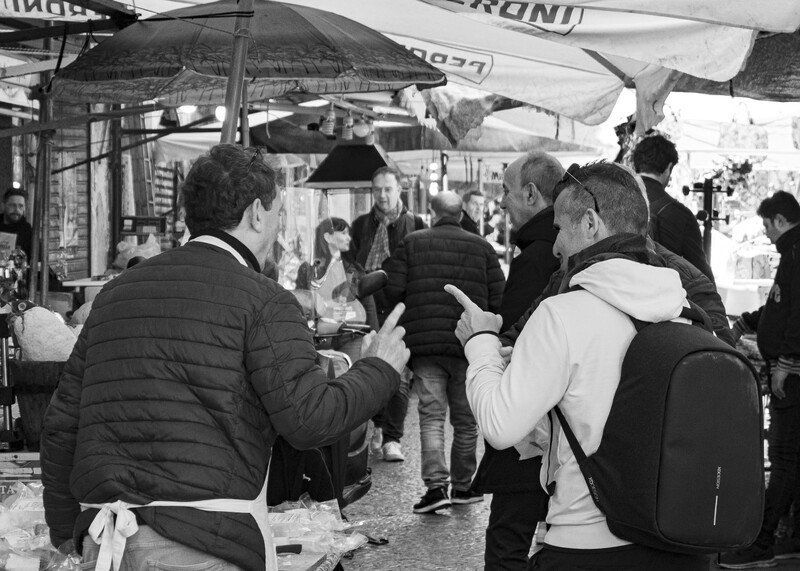Group of people gathered under umbrellas at an outdoor market or event in black and white.