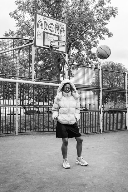 Person shooting a basketball at an outdoor court with arena signage visible behind chain-link fencing.
