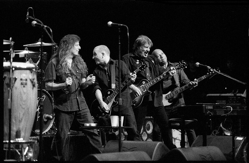 Band performing on stage with multiple guitarists, vocalist, and drummer under concert lighting.
