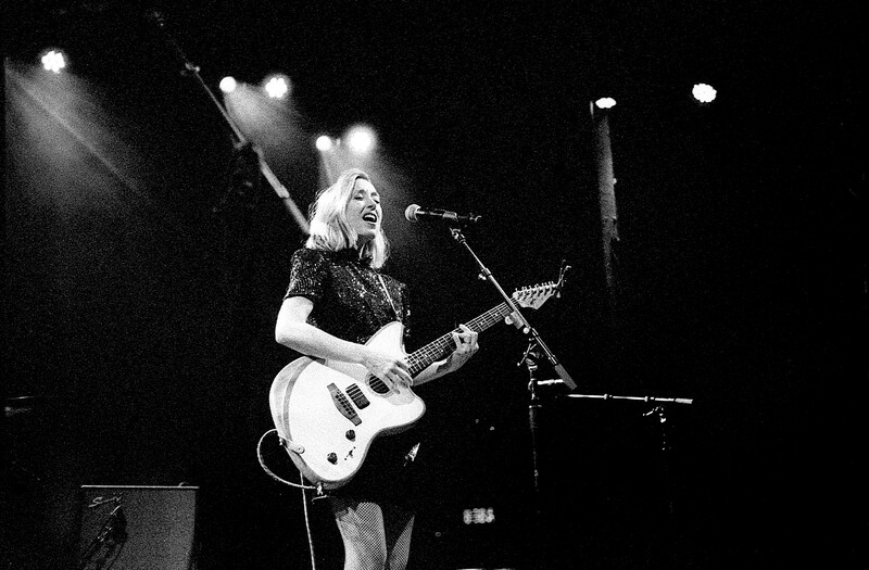 Musician performing with electric guitar under dramatic stage lighting against a dark background.