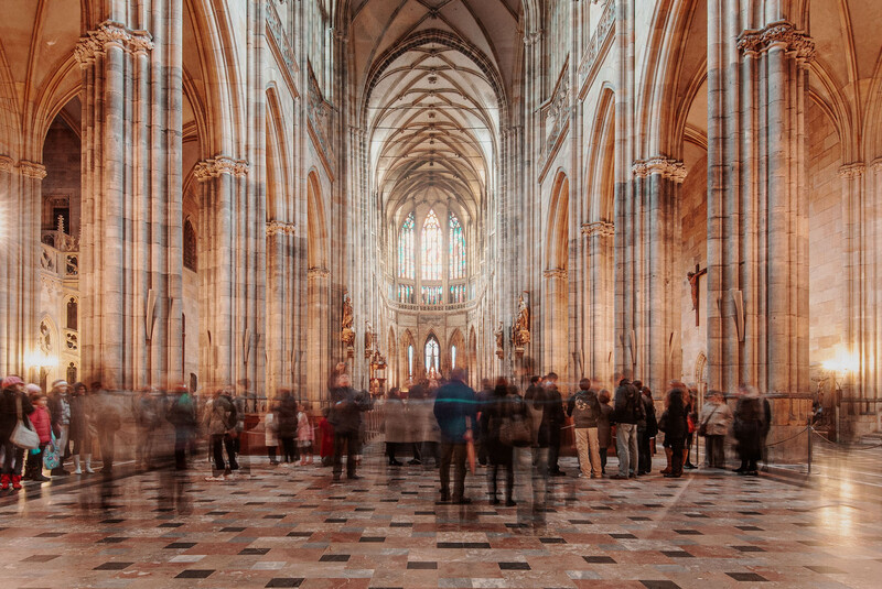 Interior of a Gothic cathedral with soaring ribbed vaults, columns, and a distant altar, populated with blurred visitors.