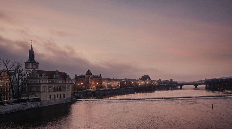 European riverside cityscape at dusk with illuminated buildings reflected in calm water under pink and purple sky.