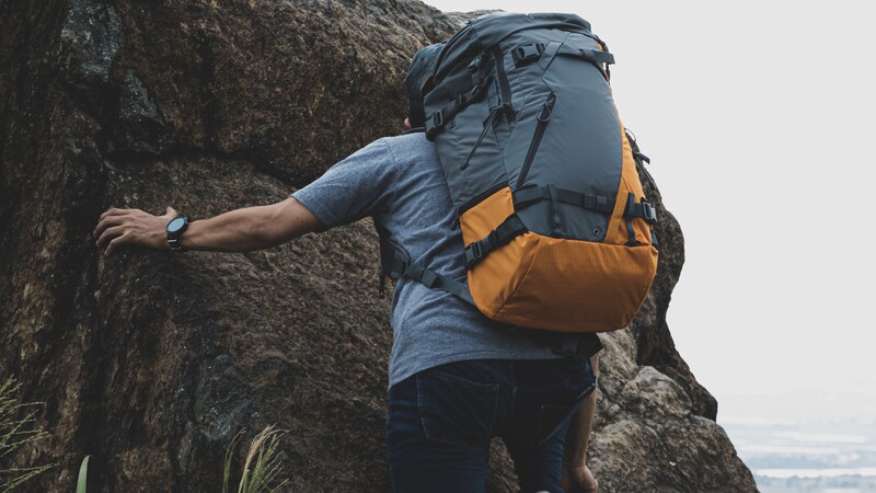 Rock climber in blue shirt and orange backpack scaling a textured stone face.