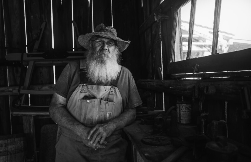Bearded man in overalls and cowboy hat standing in a rustic workshop with tools and wooden structures visible.