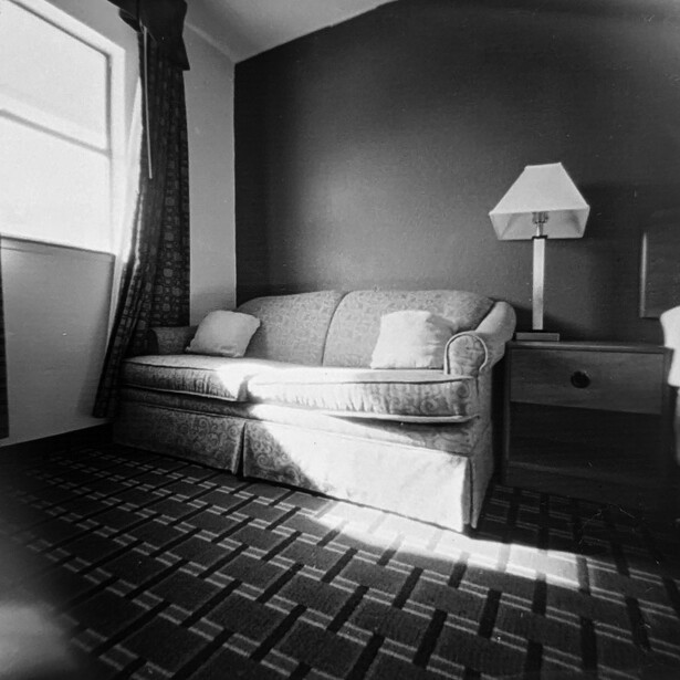 Black and white photograph of a minimalist bedroom with a daybed against a dark wall, a table lamp, and geometric floor pattern.