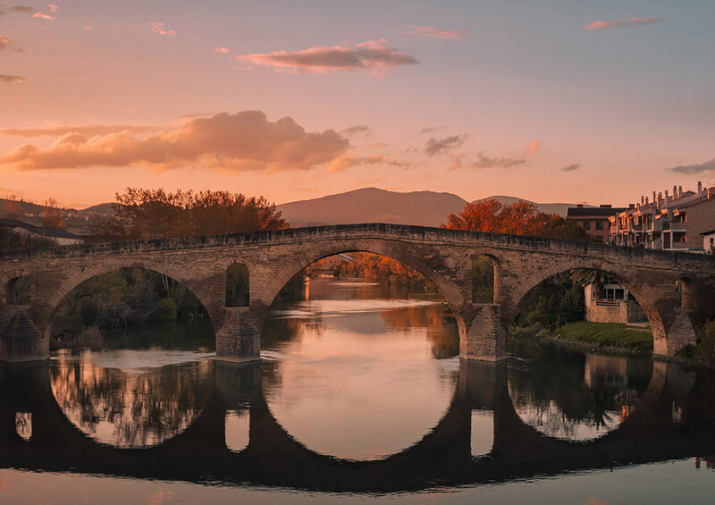 Medieval stone bridge with multiple arches reflected in still water at sunset, mountains visible in distance.