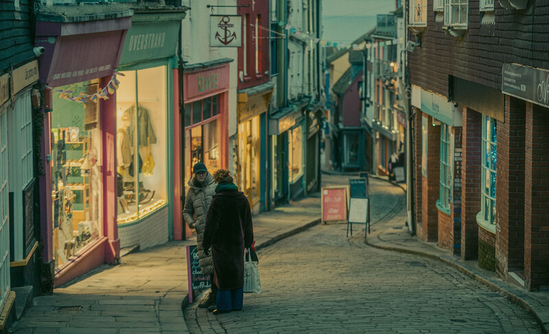 Person walking down a narrow European cobblestone street lined with illuminated shop windows at dusk.