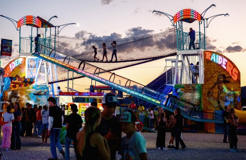 Colorful carnival ride with two tall towers and a connecting bridge structure at dusk, surrounded by crowds of visitors.