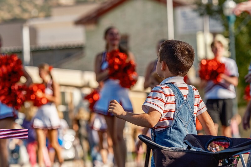 Child in striped shirt and denim vest watching Fourth of July parade with red decorations and flag visible.