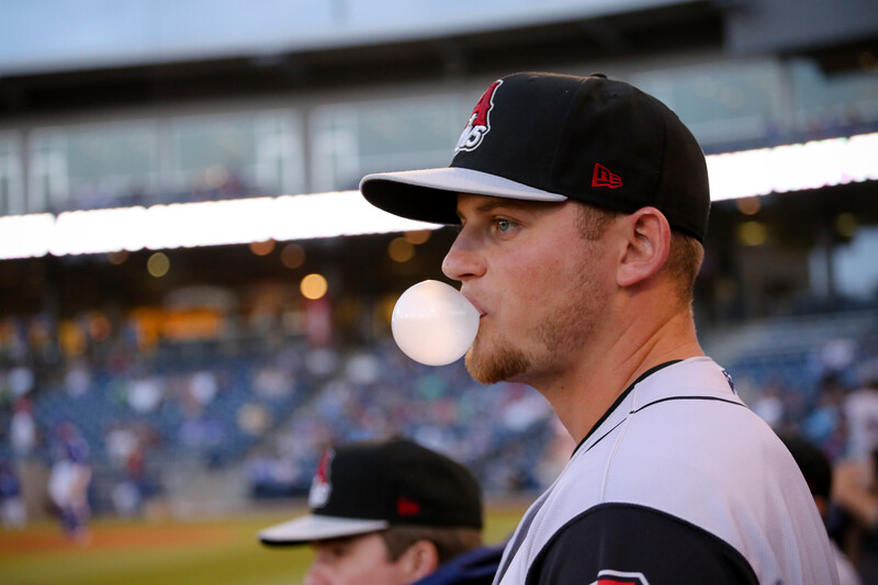 Baseball player in profile wearing a black and red cap and gray uniform, blowing a bubble with gum during a game.