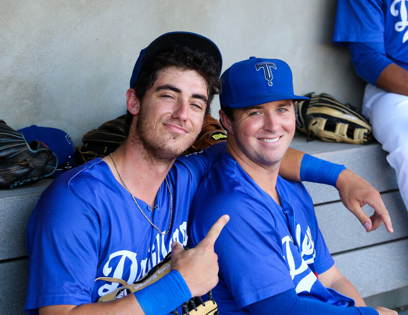 Two baseball players in blue uniforms sitting together in a dugout, smiling at the camera.