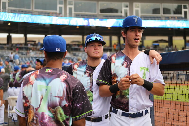 Three baseball players in blue uniforms with vibrant floral-patterned sleeves stand together at dugout railing.