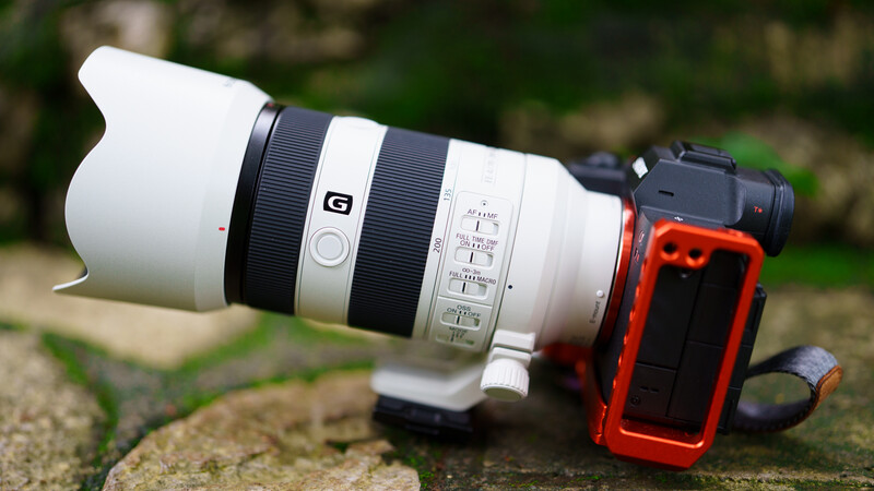 Sony G-series zoom lens with white barrel mounted on red L-bracket, positioned on sandy ground with blurred foliage background.