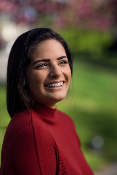 Woman in red long-sleeve shirt smiling at camera outdoors with blurred green background.