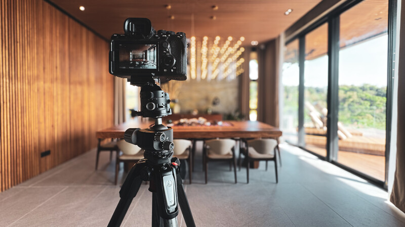 Professional camera mounted on tripod in modern dining room with floor-to-ceiling windows overlooking landscape.